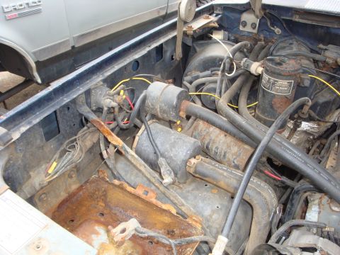 1989 Ford Bronco - Looking down into a stripped, weathered engine bay with the battery tray rusted through, showing an ignition coil, canister-style filters,…