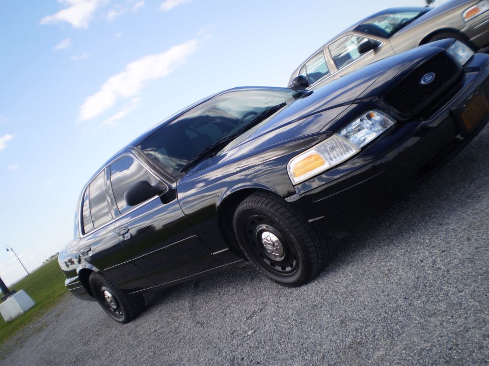 Black 2003 Ford Crown Victoria, front quarter view, parked on gravel with another car behind