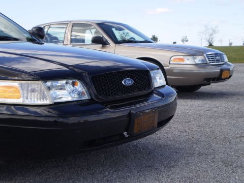 2003 Ford Crown Victoria - Two Ford Crown Victorias are parked side-by-side, with the black car in the foreground showing its grille and headlights.