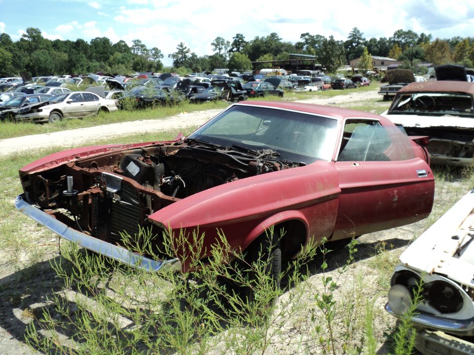 Red coupe with hood and engine removed, parked in a junkyard