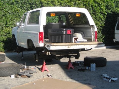 1986 Ford Bronco - White 1986 Ford Bronco is lifted on jack stands with its tailgate open, surrounded by tools and spare tires.