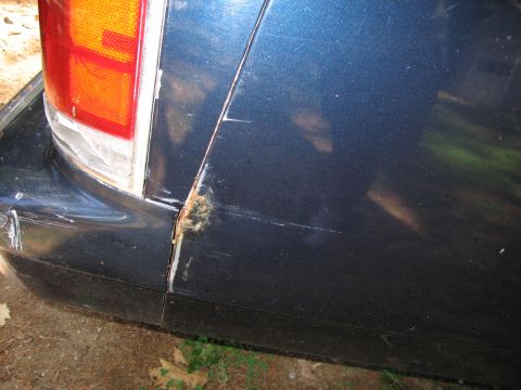 1991 Ford LTD Crown Victoria - Close-up of a dark blue car's rear quarter panel, revealing rust and damage around the seam near the taillight.