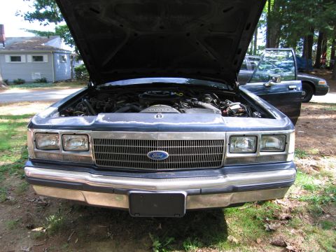 1991 Ford LTD Crown Victoria - Front view of a 1991 Ford Crown Victoria with its hood open, revealing the engine bay. Another car is visible in the background.