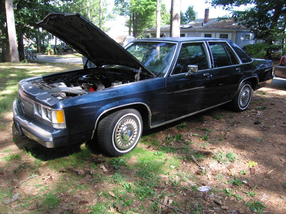 Dark blue sedan, hood open, showing engine. Whitewall tires with chrome hubcaps. Visible chrome trim along the bottom