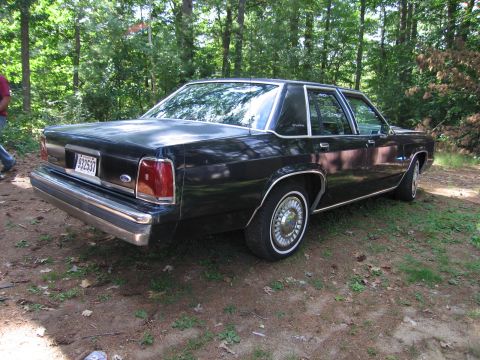1991 Ford LTD Crown Victoria - Three-quarter rear view of a black boxy Ford full-size four-door sedan parked on a dirt and grass clearing in the woods, with chrome bumper…
