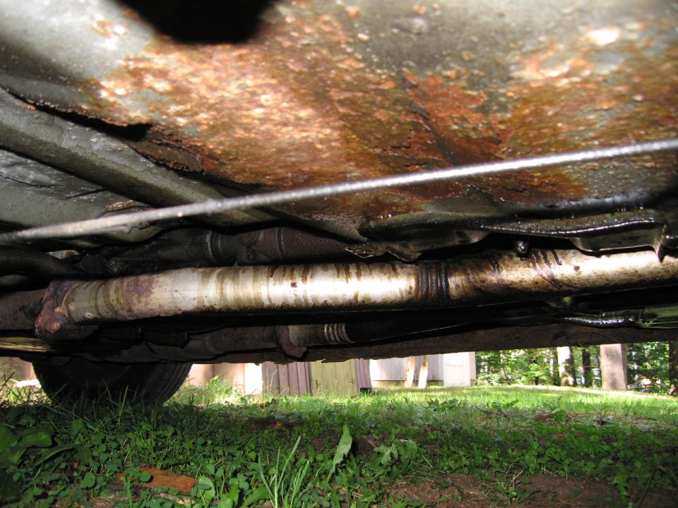Underside of car showing rusted frame and exhaust pipes. A tire is visible on the left