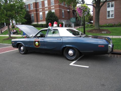 1991 Ford LTD Crown Victoria - two-tone blue Dodge police sedan with a circular agency shield and open hood parked outside a brick building.