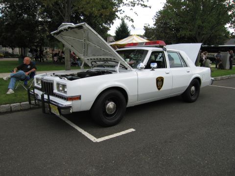 1991 Ford LTD Crown Victoria - white police sedan with its hood up showing the engine, a round door shield and roof light bar at a curb.