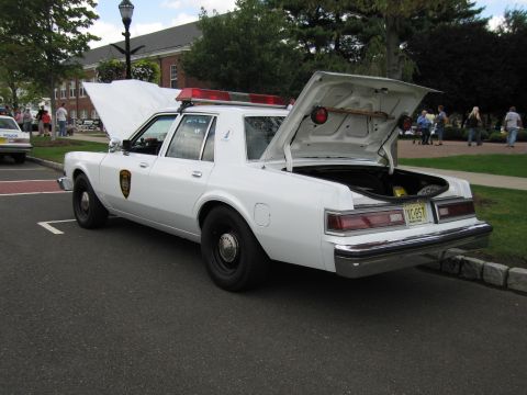 1991 Ford LTD Crown Victoria - white police sedan with open trunk and rear-deck light bar, round door shield, parked outside a building.