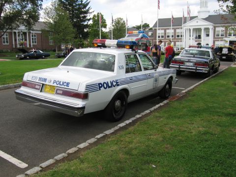 1991 Ford LTD Crown Victoria - white sedan marked 'MIAMI POLICE 839' parked at a curb during an outdoor show, with another patrol car ahead.