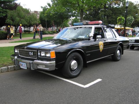 1991 Ford LTD Crown Victoria - black Chevrolet police sedan with gold star shield and roof lights, number 154, at an outdoor car show with spectators.