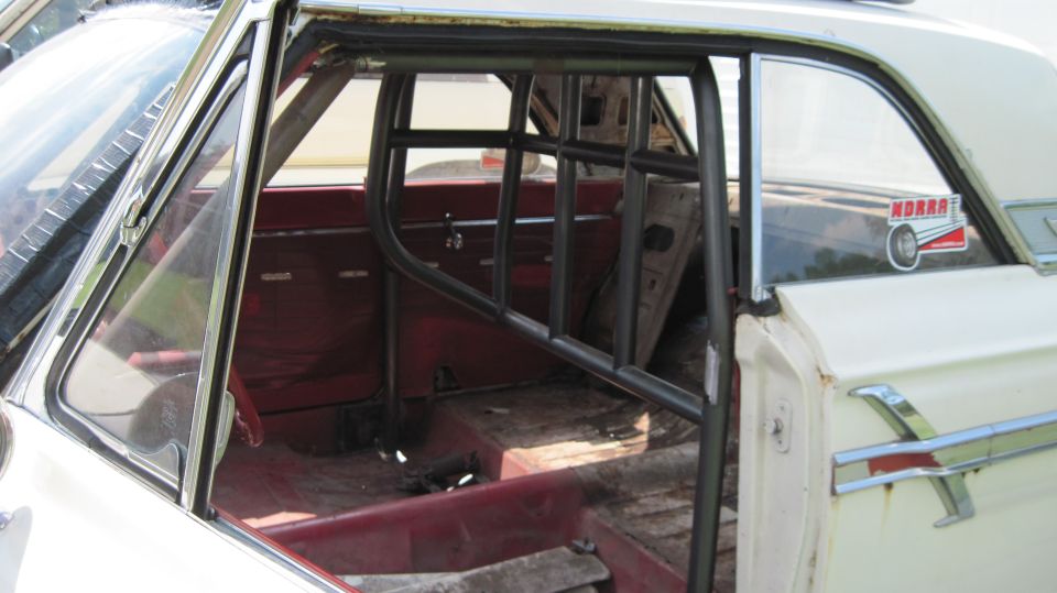 Interior of a white 1964 Ford Fairlane with a roll cage, red seats, and a Moroso sticker on the rear window