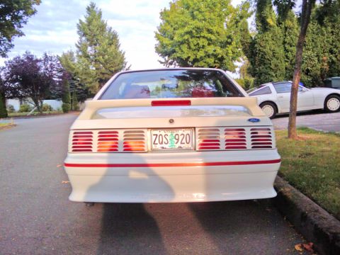 1987 Ford Mustang - Rear view of a white 1987 Ford Mustang parked on a street, with another white car visible in the background.