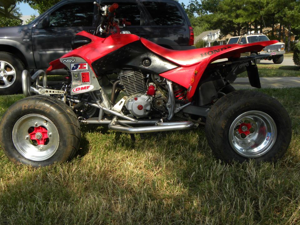 Red Honda sport ATV on grass with polished wheels, aftermarket A-arms and sponsor stickers
