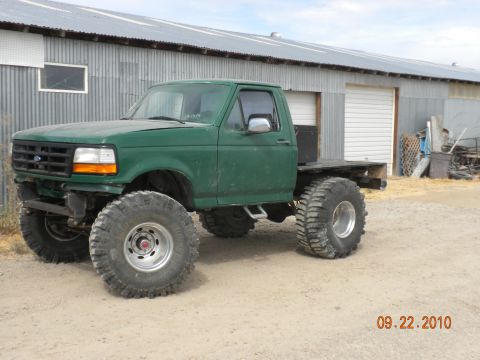 1989 Ford Bronco - This modified green flatbed truck features oversized tires and a lifted suspension, suggesting it's built for off-road use.