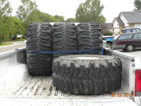 1989 Ford Bronco - Oversized tires are loaded into the bed of a pickup truck, with other vehicles and houses in the background.