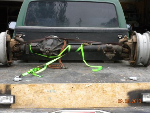 1989 Ford Bronco - A rear axle assembly, complete with wheels and secured with straps, is loaded onto the bed of a green pickup truck.