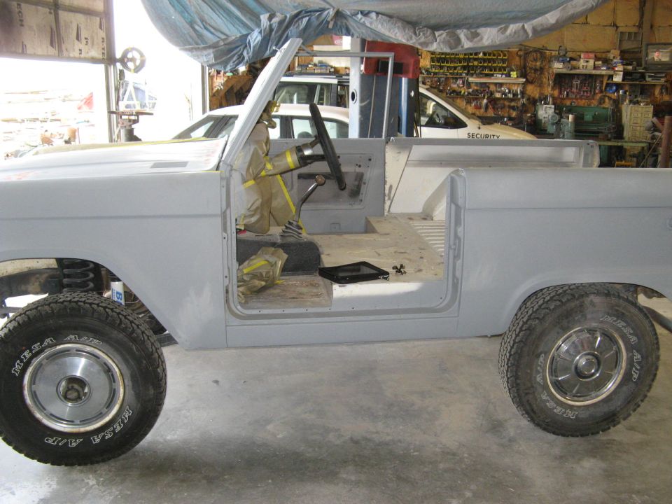 Side view of a 1966 Ford Bronco in primer, doors open, interior stripped down. Another vehicle is visible behind it