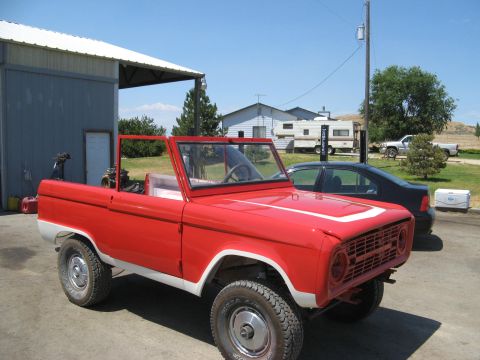 1966 Ford Bronco - A red convertible SUV is parked in front of a building with a white roof. Other vehicles and a camper are visible in the background.
