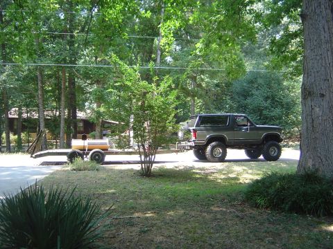 1986 Ford Bronco - Side view of a lifted dark green two-tone Ford Bronco on oversized tires and chrome wagon wheels, towing a tandem-axle flatbed trailer down…