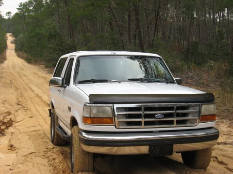 1994 Ford Bronco - White 1994 Ford Bronco navigates a sandy, off-road trail surrounded by trees.