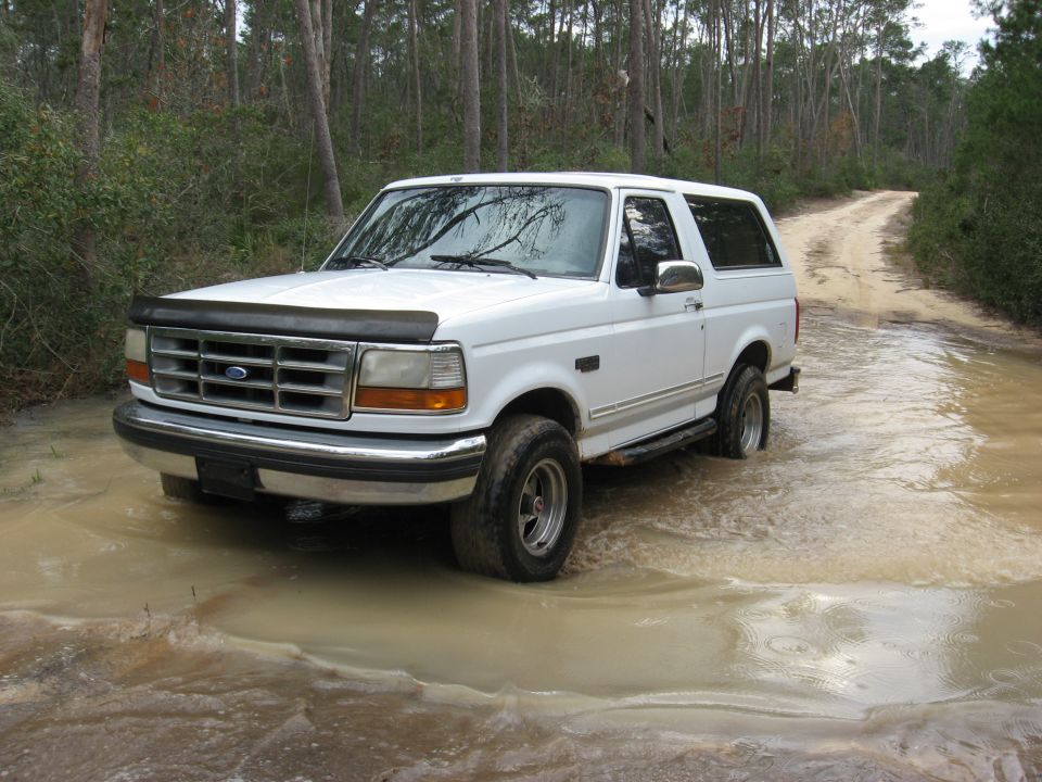 White 1994 Ford Bronco driving through a muddy trail in the woods