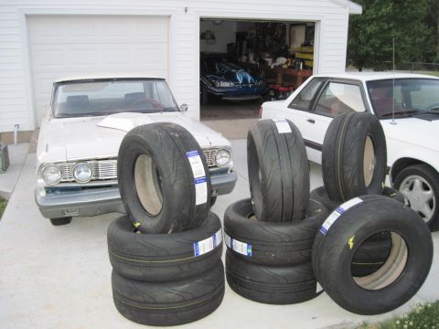 1992 Ford Mustang - A vintage Ford Falcon sits outside a garage with several performance tires stacked nearby, while another car is parked to its right.