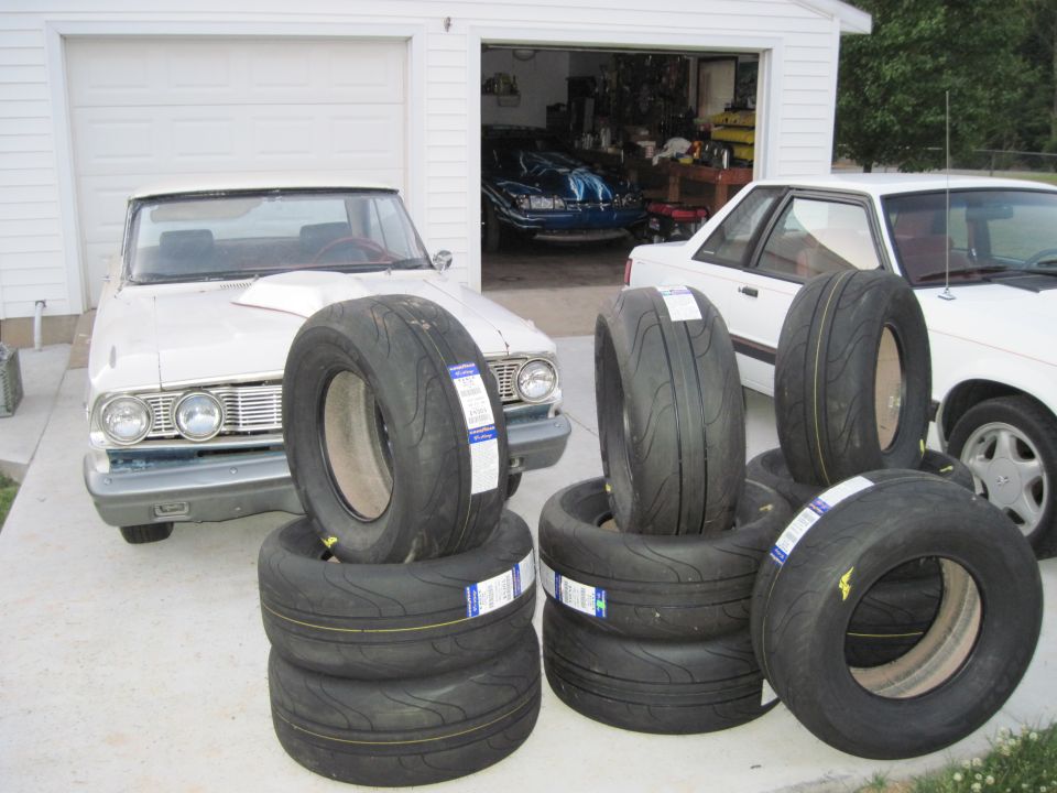 White 1964 Ford Falcon with a hood scoop and a stack of performance tires in front of a garage. Another white car is parked to the right, and a blue car is visible inside the garage