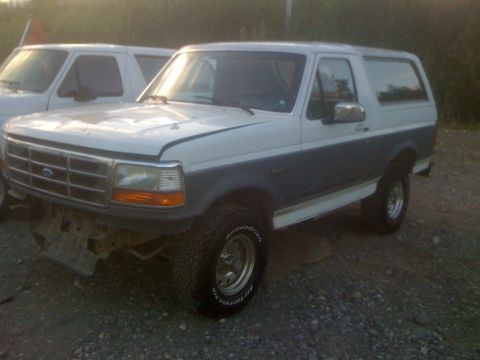 1993 Ford Bronco - A two-tone Ford Bronco is parked outdoors next to another vehicle, with damage visible on its front bumper.