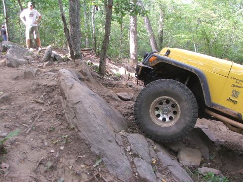 1996 Ford Bronco - A yellow Jeep Wrangler navigates a rocky trail in a wooded area, with two people visible in the background.