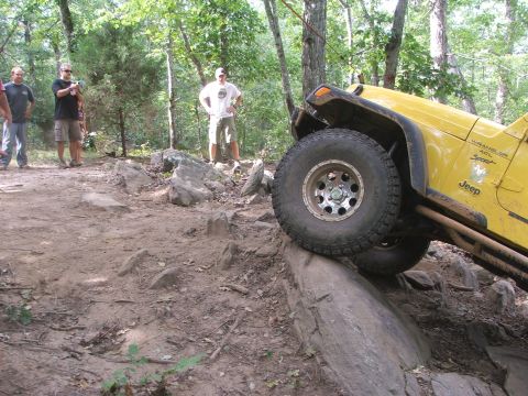 1996 Ford Bronco - A yellow Jeep Wrangler navigates a challenging rocky off-road course with spectators watching.
