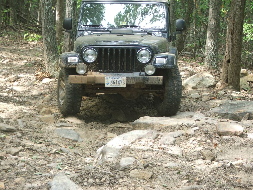 Front view of a dark green Jeep Wrangler TJ driving over rocks on a wooded trail. Virginia license plate 8614VB is visible