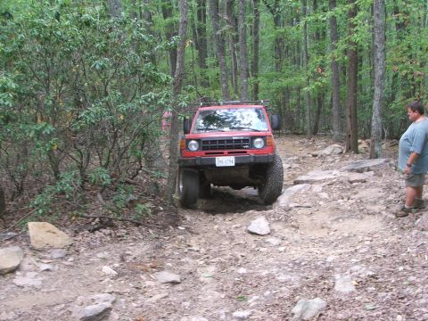 1996 Ford Bronco - A red SUV tackles a challenging, rocky path through a dense forest, with a person observing from the side.