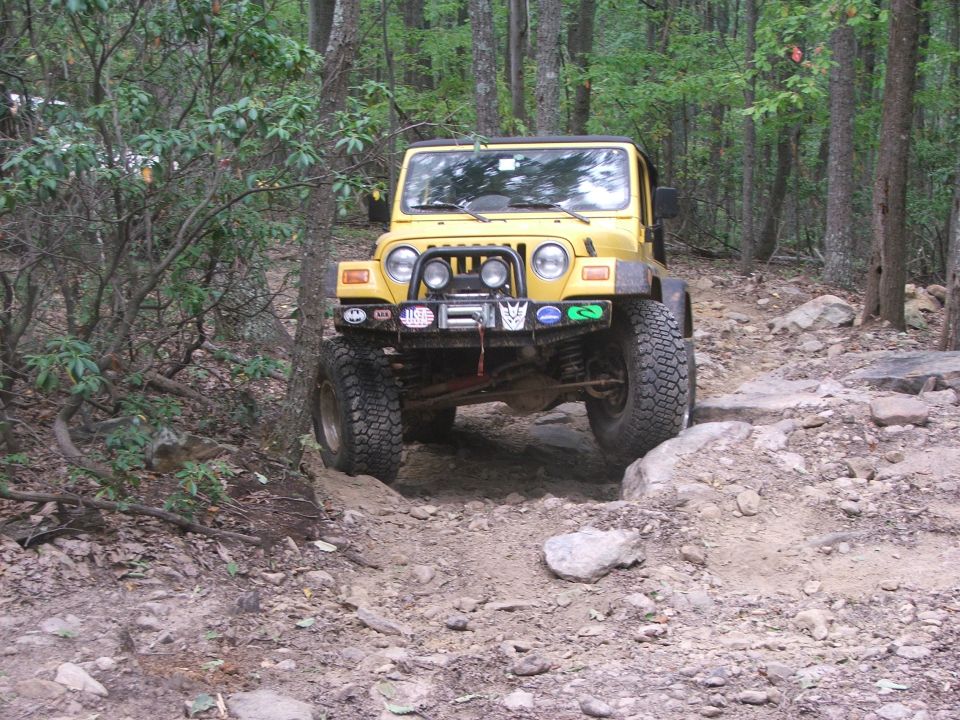 Yellow Jeep Wrangler driving over rocks on a wooded trail. It has a winch and several stickers on the front bumper