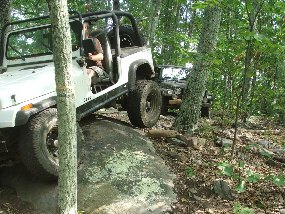 White Jeep climbing over a rock on a wooded trail, with another Jeep behind it