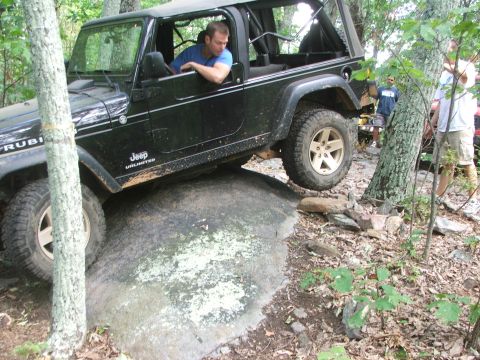 1996 Ford Bronco - A black Jeep Wrangler is shown off-roading over a large rock obstacle in a wooded area.