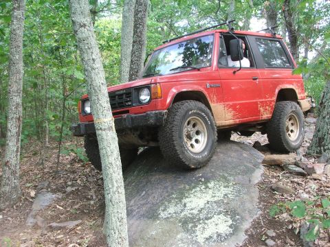 1996 Ford Bronco - This red SUV is navigating a rocky obstacle on an off-road trail surrounded by trees.