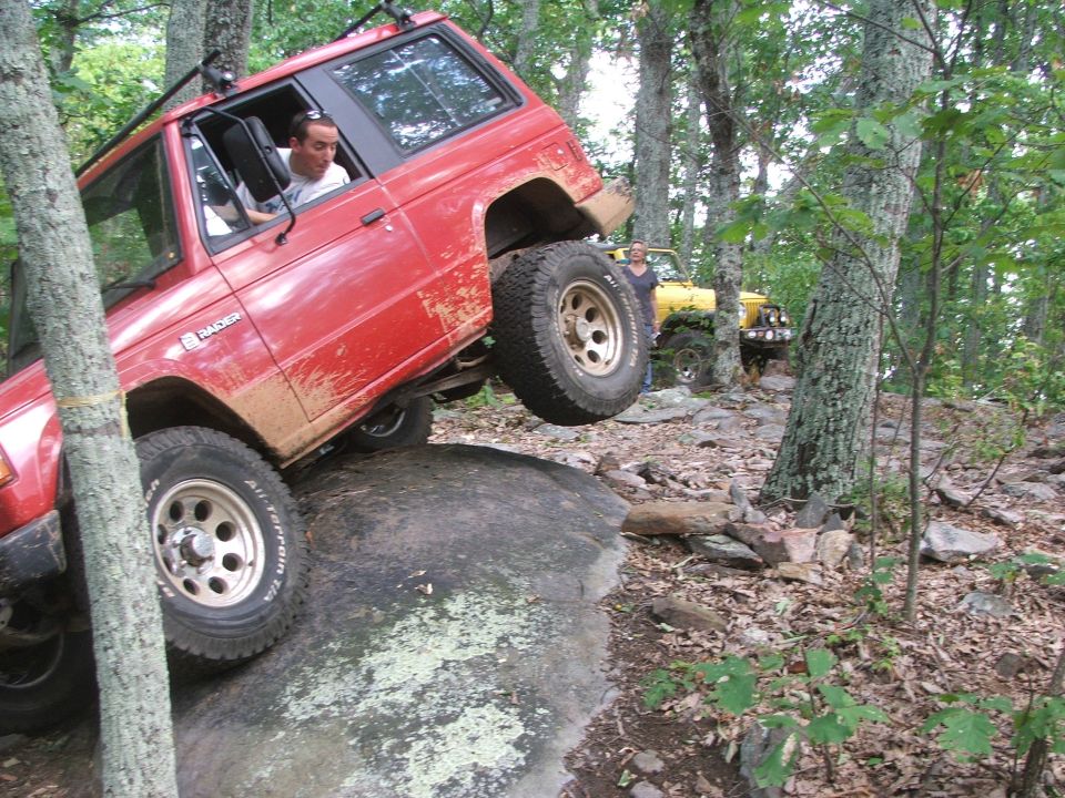 Red off-road SUV with one wheel on a large rock, yellow off-road vehicle behind it in a wooded area