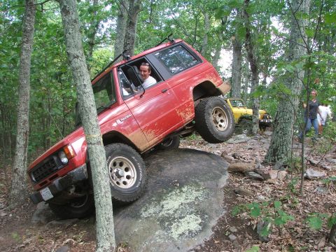 1996 Ford Bronco - An off-road vehicle navigates a challenging rocky terrain in a wooded area, with another vehicle and spectators present.