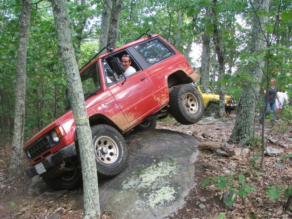 Red SUV on a rock, yellow Jeep behind it, people watching in woods