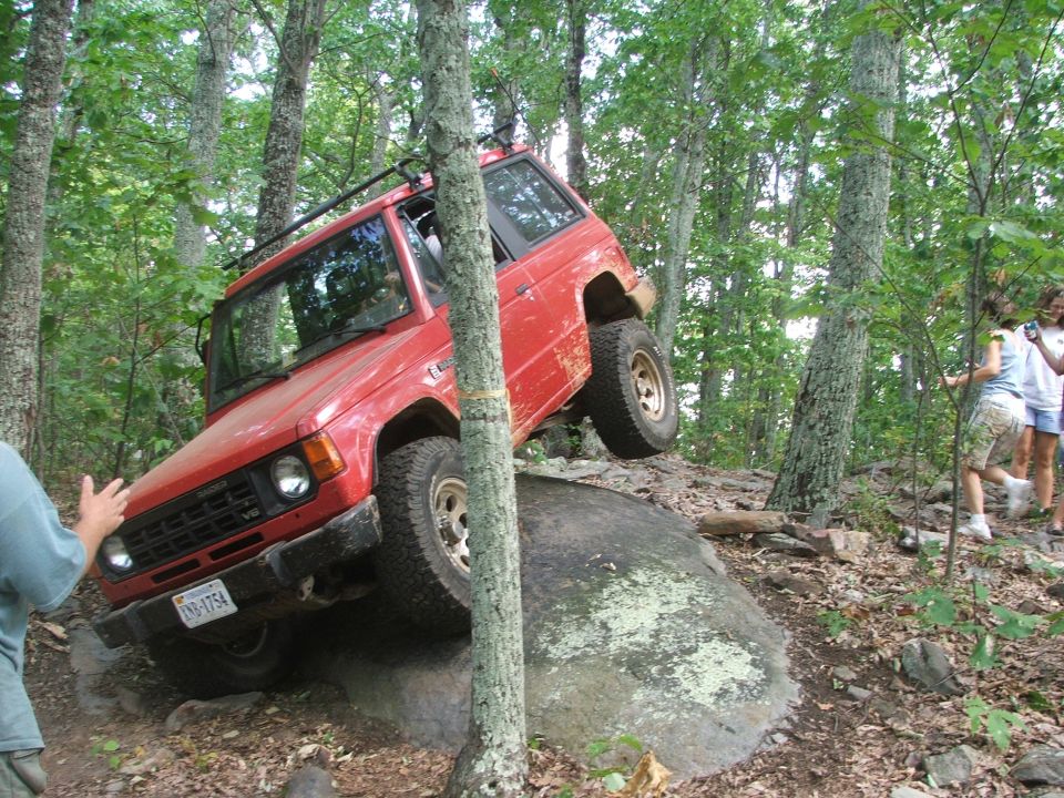 Red SUV with roof rack navigating a large rock on a wooded trail