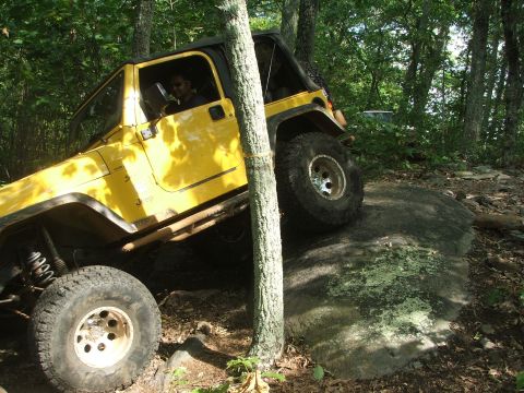 1996 Ford Bronco - A yellow Jeep Wrangler navigates a challenging rocky obstacle on a wooded trail, demonstrating its off-road capabilities.