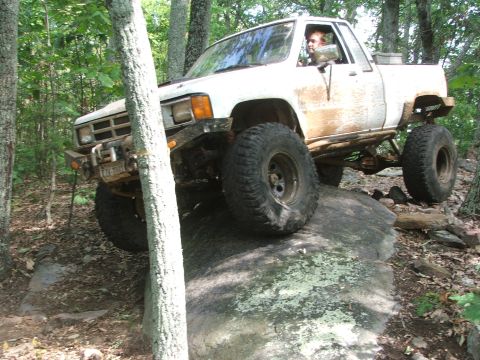 1996 Ford Bronco - White pickup truck with large off-road tires climbing over a rock in a wooded area.