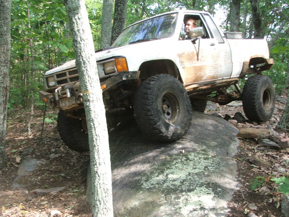 White pickup truck with large off-road tires climbing over a rock in a wooded area