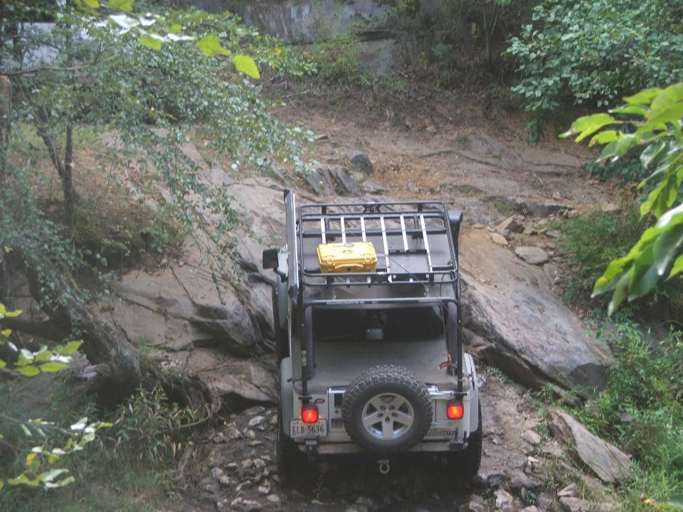 Jeep Wrangler with roof rack and yellow case driving on a rocky, wooded trail. License plate reads KLR-5636