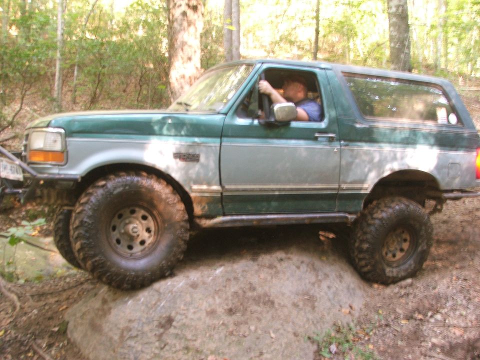 Green Ford Bronco driving over a rock on a wooded trail. Driver visible
