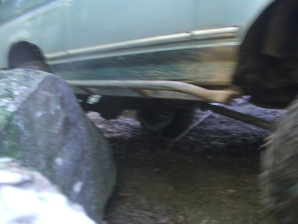 Green vehicle undercarriage with exposed frame, exhaust pipe, and tires on a dirt surface