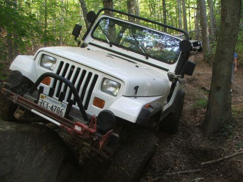 1996 Ford Bronco - A white Jeep Wrangler navigates a rocky obstacle on a wooded trail, with another vehicle in the background.
