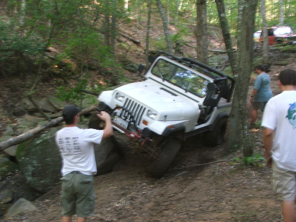 White Jeep Wrangler on a dirt trail, front-left tire angled up a rock, with two men standing nearby