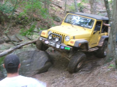 1996 Ford Bronco - A yellow Jeep Wrangler tackles a challenging off-road course with large rocks and trees.